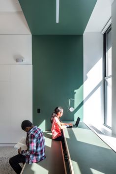 two people sitting on benches in a room with green walls and large windows, looking at their laptops