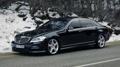 a black mercedes s - class parked on the side of a road in front of snow covered trees