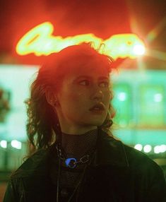 a woman standing in front of a neon sign at night with her hair blowing in the wind