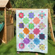 a person holding up a colorful quilt on the grass in front of a wooden structure