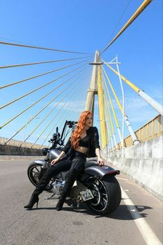 a woman sitting on top of a motorcycle in front of a suspension bridge over water