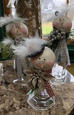 three snowmen are sitting in glass vases on a marble counter top, decorated with silver and white feathers