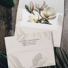 two envelopes with white flowers on them sitting next to green leaves and wood planks