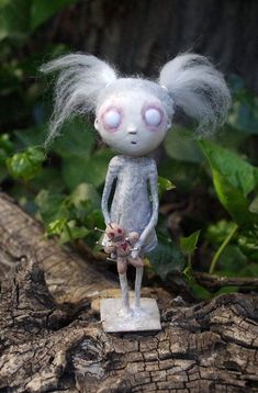 a small doll with white hair standing on a tree stump in front of green leaves