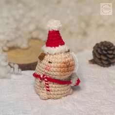 a small crocheted snowman sitting in the snow next to a pine cone