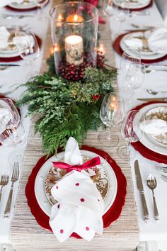 the table is set for christmas dinner with red and white plates, silverware, candles and greenery