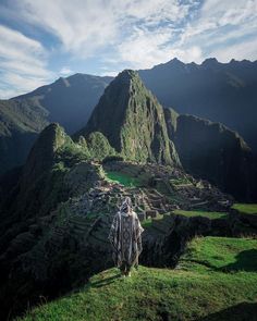 a man standing on top of a lush green hillside next to a large mountain range