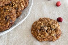 some cookies and cherries are on a white tablecloth next to a bowl of cranberry oatmeal cookies