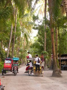 three people riding on the back of a motorcycle down a dirt road surrounded by palm trees