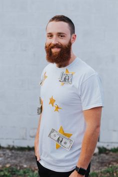 a man with a beard standing in front of a white wall wearing money on his shirt