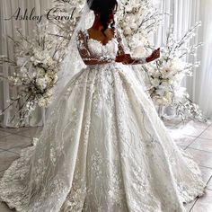 a woman standing in front of a white backdrop wearing a wedding dress with long sleeves