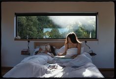 a woman sitting in bed reading a book and drinking coffee while looking out the window