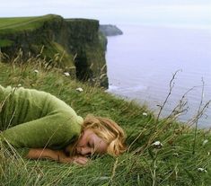 a woman laying on top of a lush green hillside next to the ocean with her eyes closed