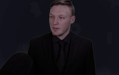 a man in a black suit and tie is talking to the camera while holding a microphone