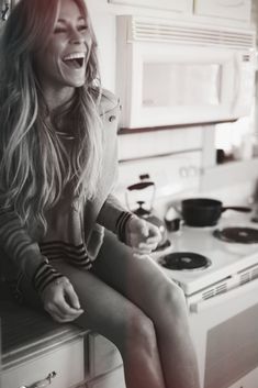 a woman sitting on top of a kitchen counter next to a stove and oven smiling