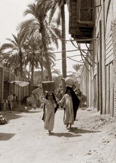 two women are walking down the street holding hands and carrying boxes on their heads, with palm trees in the background