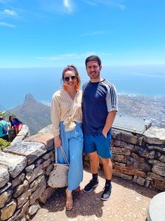 a man and woman standing on top of a stone wall next to each other with the ocean in the background