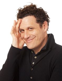 a man with curly hair and black shirt smiling at the camera while holding his hand to his head