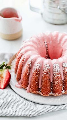 a bundt cake with pink icing on a white plate next to a strawberry