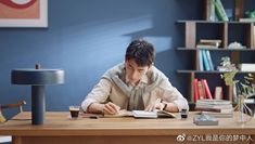 a young man sitting at a desk writing in an open book and holding a pen