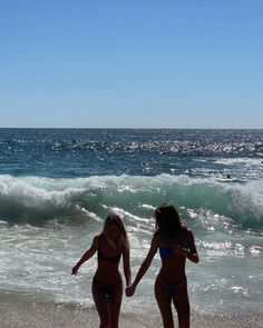 two women in bikinis standing on the beach next to the ocean and holding hands