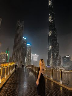 a woman standing on top of a bridge in front of some tall buildings at night