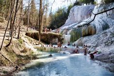 many people are swimming in the water near some rocks and trees, while others stand around