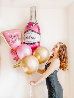 a woman in a dress holding balloons and a bottle of champagne next to a wall