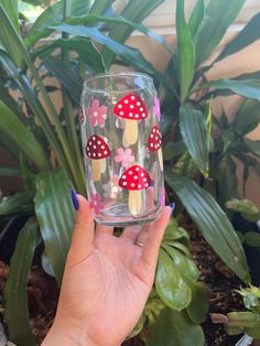 a hand holding a glass with mushrooms painted on it in front of a potted plant