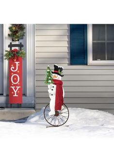 a snowman on a bicycle in front of a house with a joy sign behind it