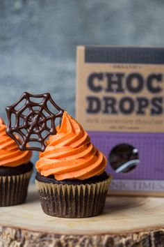 two chocolate cupcakes with orange frosting and spider web decorations on top, next to a box of choc drops