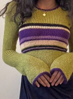 a woman wearing a green and purple sweater with her hands in her pockets while standing next to a white wall