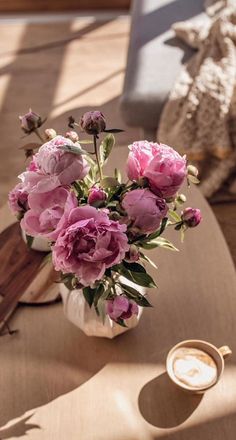 pink flowers in a white vase on a table with a cup of coffee next to it