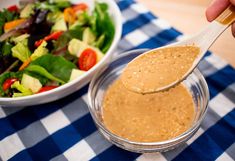 a person holding a spoon over a salad in a bowl with dressing on the side