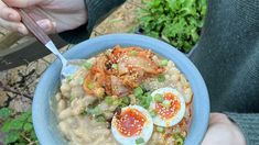 a person holding a bowl of food with eggs and vegetables in it, while another hand holds a spoon