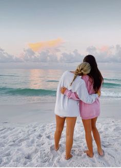 two girls standing on the beach hugging each other and looking out at the ocean while the sun is setting