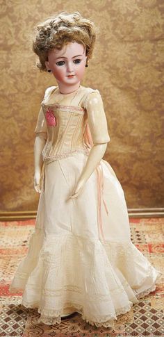 an antique porcelain doll wearing a white dress and pink flower in her hair, standing on a rug