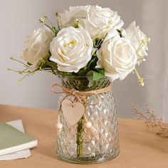 white roses in a clear glass vase with a heart shaped tag on the handle, sitting on a wooden table