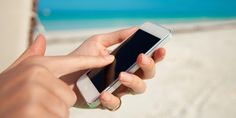 a person holding a cell phone in their hand on the beach with blue sky and white sand