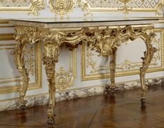 an ornate gold console table with glass top in front of a white wall and wood floor