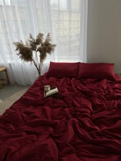 an unmade bed with red sheets and pillows in front of a window next to a plant