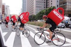 a group of people riding bikes down a street with red bags on their back's backs