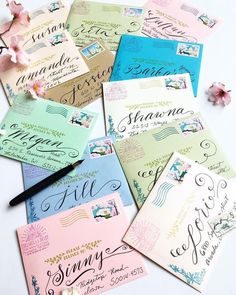 several different colored envelopes with writing on them next to a pen and flower petals