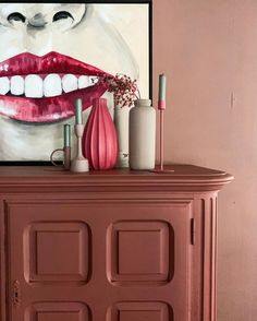 a pink cabinet with lipstick, vases and bottles on it in front of a painting