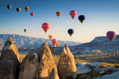 many hot air balloons flying in the sky over some rocks and mountains, with snow capped mountains in the background