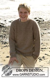 a young man standing on top of a sandy beach next to the ocean wearing a brown sweater and khaki pants