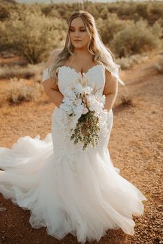 a woman in a wedding dress standing on the side of a dirt road holding a bouquet