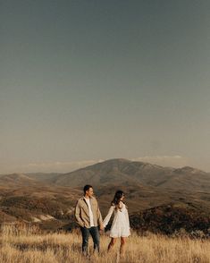 a man and woman holding hands while standing on top of a grass covered field with mountains in the background