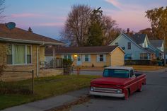 an old red truck parked in front of a yellow house at dusk with the sun going down