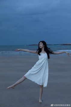 a woman in a white dress is standing on the beach with her arms spread out